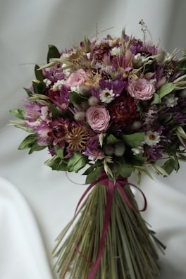A bouquet of dried flowers featuring a variety of textures and colors, including soft pink roses, vibrant purple blooms, small white flowers, and green leaves. The stems are elegantly tied with a dark pink ribbon.