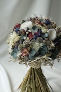 A bouquet of dried flowers beautifully arranged with a mix of various textures and colors. The arrangement includes roses, thistles, eucalyptus leaves, and other botanical elements. The bouquet displays a natural and rustic aesthetic, with each component adding to its intricate design.