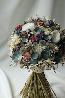 A bouquet of dried flowers beautifully arranged with a mix of various textures and colors. The arrangement includes roses, thistles, eucalyptus leaves, and other botanical elements. The bouquet displays a natural and rustic aesthetic, with each component adding to its intricate design.
