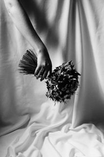 A black and white photograph showing a hand holding a bouquet of flowers against a soft, draped fabric background. The composition highlights the texture of the flowers and the drapery, creating an elegant and artistic scene.
