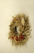 An elegant wreath made of dried flowers and grasses is displayed against a neutral background. The arrangement includes various textures and warm colors, showcasing an assortment of dried petals and stems along with clusters of brown and beige elements.