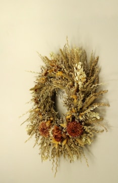 An elegant wreath made of dried flowers and grasses is displayed against a neutral background. The arrangement includes various textures and warm colors, showcasing an assortment of dried petals and stems along with clusters of brown and beige elements.