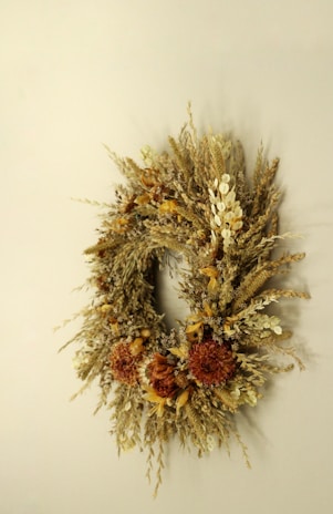 An elegant wreath made of dried flowers and grasses is displayed against a neutral background. The arrangement includes various textures and warm colors, showcasing an assortment of dried petals and stems along with clusters of brown and beige elements.