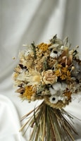 A beautifully arranged bouquet of dried flowers featuring a mix of textures and colors, including yellow, white, and beige blooms with greenery. The bouquet is set against a soft, cream-colored backdrop, giving it an elegant and rustic appearance.