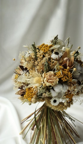 A beautifully arranged bouquet of dried flowers featuring a mix of textures and colors, including yellow, white, and beige blooms with greenery. The bouquet is set against a soft, cream-colored backdrop, giving it an elegant and rustic appearance.