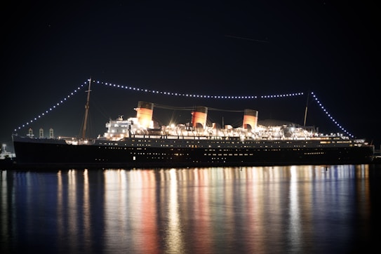 A large illuminated ocean liner with three distinct smokestacks casts a glowing reflection on the calm water at night. Strings of lights drape over the ship, enhancing its grandeur and outlining its structure against the dark sky. The ship appears stationary, with its numerous windows and decks clearly visible, giving an impression of elegance and nostalgia.