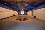 A spacious room with a high, blue ceiling featuring wooden beams. The walls are adorned with earthy installations and sculptures. The floor has circular art pieces with various natural elements. The ambient lighting is subtle, creating a calm and focused atmosphere. The room extends to a projection or screen at the far end.