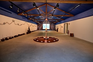 A spacious room with a high, blue ceiling featuring wooden beams. The walls are adorned with earthy installations and sculptures. The floor has circular art pieces with various natural elements. The ambient lighting is subtle, creating a calm and focused atmosphere. The room extends to a projection or screen at the far end.