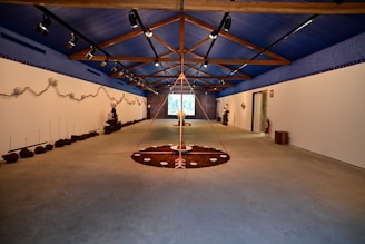 A spacious room with a high, blue ceiling featuring wooden beams. The walls are adorned with earthy installations and sculptures. The floor has circular art pieces with various natural elements. The ambient lighting is subtle, creating a calm and focused atmosphere. The room extends to a projection or screen at the far end.