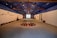 A spacious room with a high, blue ceiling featuring wooden beams. The walls are adorned with earthy installations and sculptures. The floor has circular art pieces with various natural elements. The ambient lighting is subtle, creating a calm and focused atmosphere. The room extends to a projection or screen at the far end.
