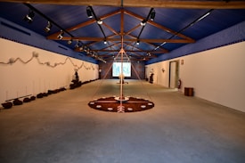 A spacious room with a high, blue ceiling featuring wooden beams. The walls are adorned with earthy installations and sculptures. The floor has circular art pieces with various natural elements. The ambient lighting is subtle, creating a calm and focused atmosphere. The room extends to a projection or screen at the far end.