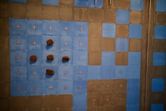 A wall installation composed of blue and brown squares arranged in a grid pattern. The blue squares are embellished with small crosses and natural rock formations attached at various points. The brown squares provide contrast, creating a checkered effect.