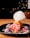 A vibrant dessert featuring a cylindrical pastry filled with pink ice cream on a black plate. Cotton candy with colorful sprinkles is perched on top, and the plate is decorated with strawberries, purple meringues, and scattered sprinkles.