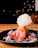 A vibrant dessert featuring a cylindrical pastry filled with pink ice cream on a black plate. Cotton candy with colorful sprinkles is perched on top, and the plate is decorated with strawberries, purple meringues, and scattered sprinkles.