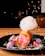 A vibrant dessert featuring a cylindrical pastry filled with pink ice cream on a black plate. Cotton candy with colorful sprinkles is perched on top, and the plate is decorated with strawberries, purple meringues, and scattered sprinkles.