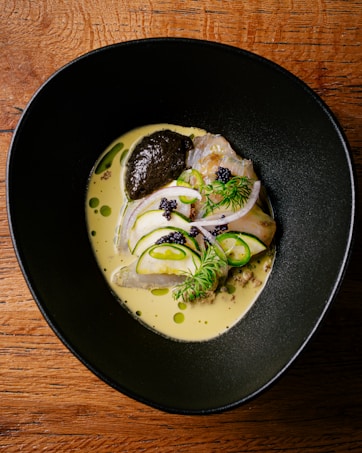 A sophisticated dish presented on a textured wooden table. The dish features slices of fish, garnished with dill, cucumber slices, red onion, and dark-colored roe. It rests in a creamy yellow sauce with a dollop of dark sauce or paste on the side.