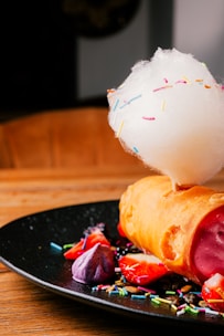 A creative dessert display features a rolled pastry filled with a red or pink creamy filling, topped with a cloud of cotton candy sprinkled with colorful decorations. Surrounding the pastry are sliced strawberries, a purple meringue, and a variety of vibrant sprinkles scattered on a black plate. The dish is set on a wooden table.
