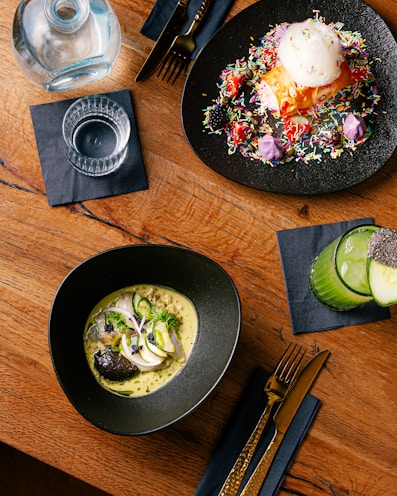 A wooden table with a modern culinary spread, including two elegant dishes, a drink, and cutlery. One dish appears to be a colorful dessert with sprinkles and a scoop of ice cream on a black plate. Another dish contains a gourmet preparation with fish, garnished with herbs and slices of vegetables, presented in a black bowl. A glass of green juice garnished with chia seeds is placed on the side next to gold-colored cutlery on a dark napkin.
