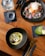 A wooden table with a modern culinary spread, including two elegant dishes, a drink, and cutlery. One dish appears to be a colorful dessert with sprinkles and a scoop of ice cream on a black plate. Another dish contains a gourmet preparation with fish, garnished with herbs and slices of vegetables, presented in a black bowl. A glass of green juice garnished with chia seeds is placed on the side next to gold-colored cutlery on a dark napkin.