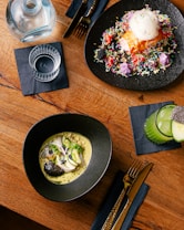 A wooden table with a modern culinary spread, including two elegant dishes, a drink, and cutlery. One dish appears to be a colorful dessert with sprinkles and a scoop of ice cream on a black plate. Another dish contains a gourmet preparation with fish, garnished with herbs and slices of vegetables, presented in a black bowl. A glass of green juice garnished with chia seeds is placed on the side next to gold-colored cutlery on a dark napkin.