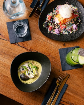 A wooden table with a modern culinary spread, including two elegant dishes, a drink, and cutlery. One dish appears to be a colorful dessert with sprinkles and a scoop of ice cream on a black plate. Another dish contains a gourmet preparation with fish, garnished with herbs and slices of vegetables, presented in a black bowl. A glass of green juice garnished with chia seeds is placed on the side next to gold-colored cutlery on a dark napkin.
