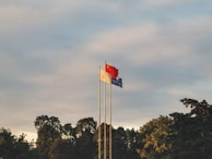 Three flags are raised on poles against a backdrop of a partly cloudy sky. The flags include the national flag of China, prominently red with yellow stars, and two other flags in white and blue. They are surrounded by the silhouette of lush green trees.