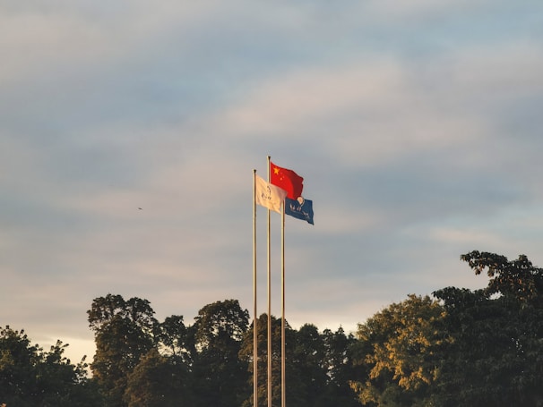 Three flags are raised on poles against a backdrop of a partly cloudy sky. The flags include the national flag of China, prominently red with yellow stars, and two other flags in white and blue. They are surrounded by the silhouette of lush green trees.
