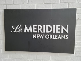 A rectangular black sign with white text displaying 'Le Meridien New Orleans' is mounted on a white brick wall.