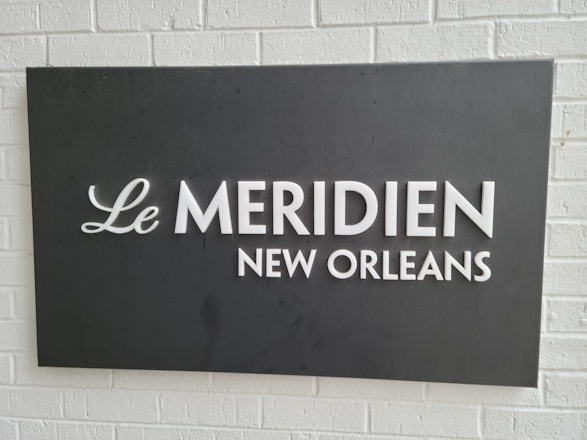 A rectangular black sign with white text displaying 'Le Meridien New Orleans' is mounted on a white brick wall.