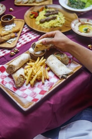 A wooden tray on a red checkered cloth holds rolled flatbreads and crispy french fries. A hand is dipping one of the flatbreads into a small bowl of white sauce. Other dishes in the background include skewered meat with rice on a round wooden platter and small plates of appetizers.