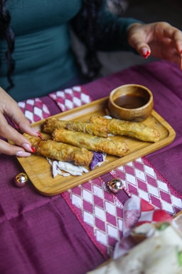 A wooden tray holds several fried rolls placed on a bed of shredded cabbage. Beside the rolls is a small wooden bowl filled with sauce. A person with red fingernails is reaching towards the tray on a purple and white patterned tablecloth, which is decorated with small metallic balls.