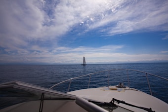 A yacht is sailing on a calm sea with a cloudy sky overhead. The perspective is from another boat, providing a view of the open water and a distant horizon.