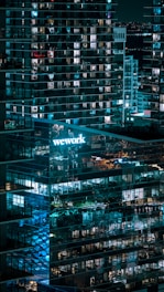 A nighttime cityscape featuring a modern glass office building with lit windows. The 'WeWork' logo is prominently displayed, casting a blue glow. Reflections of lights and other buildings are visible on the glass facade, contributing to an urban ambiance.