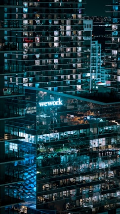 A nighttime cityscape featuring a modern glass office building with lit windows. The 'WeWork' logo is prominently displayed, casting a blue glow. Reflections of lights and other buildings are visible on the glass facade, contributing to an urban ambiance.