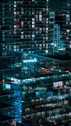 A nighttime cityscape featuring a modern glass office building with lit windows. The 'WeWork' logo is prominently displayed, casting a blue glow. Reflections of lights and other buildings are visible on the glass facade, contributing to an urban ambiance.