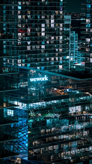 A nighttime cityscape featuring a modern glass office building with lit windows. The 'WeWork' logo is prominently displayed, casting a blue glow. Reflections of lights and other buildings are visible on the glass facade, contributing to an urban ambiance.