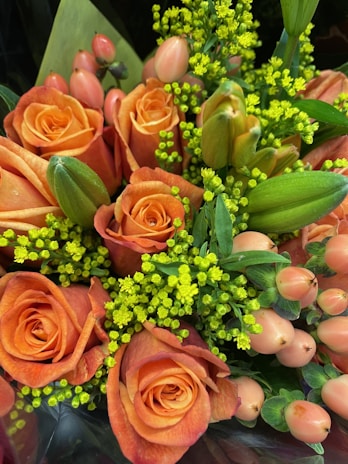 A beautifully arranged bouquet featuring vibrant orange roses, green buds, pink berries, and yellow filler flowers surrounded by lush green leaves.