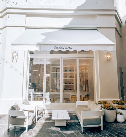 The image depicts the exterior of a boutique with a white awning that has 'la la land' written on it. In front of the store, there is a seating area with two white armchairs, a white coffee table, and a white bench. Inside the store, various items of clothing and accessories are neatly displayed on shelves. Potted flowers in white pots are arranged on the right side of the entrance.