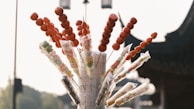 A collection of colorful candies on sticks, with a focus on red candied fruit skewers. The candies are wrapped in clear plastic and displayed like a bouquet. The background has traditional architecture with a blurred, serene setting.