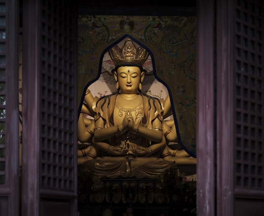 A golden statue depicting a serene deity with multiple arms is positioned inside an ornate chamber, framed by wooden doors. The setting is richly decorated, with intricate patterns and a sense of reverence.