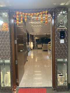 An intricately decorated entryway with a garland of marigold and yellow flowers hanging above a wooden door. The inside view reveals a well-lit interior with glossy marble flooring, modern ceiling designs, and contemporary furniture. Decorative items and soft lighting add to the opulent ambiance.