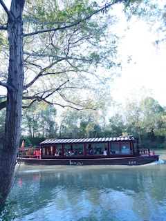 A traditional wooden boat with a curved roof is floating on a calm river. The boat is carrying several passengers and is enclosed with windows. Tall trees with lush green leaves line the riverbank, creating a serene and natural backdrop.