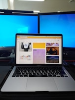 A workspace setup featuring a laptop displaying a webpage with various images, flanked by two large monitors with blue desktop backgrounds. The laptop is positioned on a dark desk with a keyboard and other items partially visible.