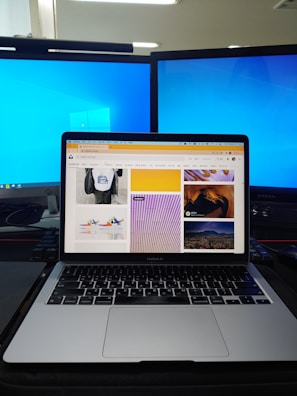 A workspace setup featuring a laptop displaying a webpage with various images, flanked by two large monitors with blue desktop backgrounds. The laptop is positioned on a dark desk with a keyboard and other items partially visible.