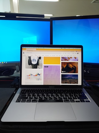 A workspace setup featuring a laptop displaying a webpage with various images, flanked by two large monitors with blue desktop backgrounds. The laptop is positioned on a dark desk with a keyboard and other items partially visible.