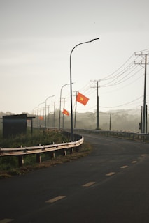 A curved road with a metal guardrail on the left side, adorned with several red flags featuring a yellow star. Tall streetlights align the road, and power lines run overhead. The background appears hazy, suggesting early morning or foggy conditions, with some vegetation visible near the roadside.