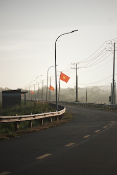 A curved road with a metal guardrail on the left side, adorned with several red flags featuring a yellow star. Tall streetlights align the road, and power lines run overhead. The background appears hazy, suggesting early morning or foggy conditions, with some vegetation visible near the roadside.