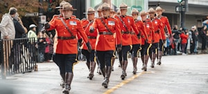 A line of uniformed officers wearing traditional red coats and brown hats march in step during a parade. The officers are accessorized with dark blue pants featuring a yellow stripe, brown boots, and belts. Medals can be seen on their chests. Spectators line the street, some taking photos, and a few trees and buildings are visible in the background.