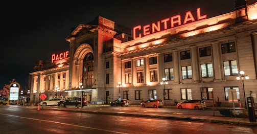 A grand, historic-looking building illuminated by warm, glowing lights at night. Red neon letters spell out 'Pacific Central' on the roof. The building features a classic architectural style with columns and numerous windows. In front, several parked cars and taxis are visible on the street, along with some street lamps casting light on the wet pavement.
