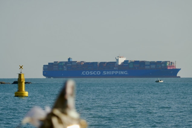 A large container ship labeled COSCO SHIPPING is prominently positioned on the ocean, with a small yellow buoy floating on the left side. A small boat is visible in the foreground, and the overall scene is captured under a clear sky.